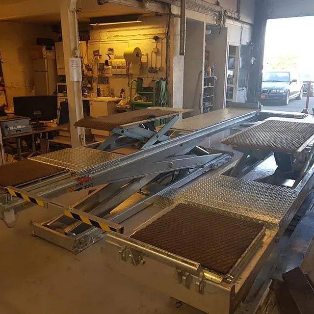 Hevet løftebukk i verksted, omgitt av arbeidsbenker, kontorhjørne og åpning mot uteområde.